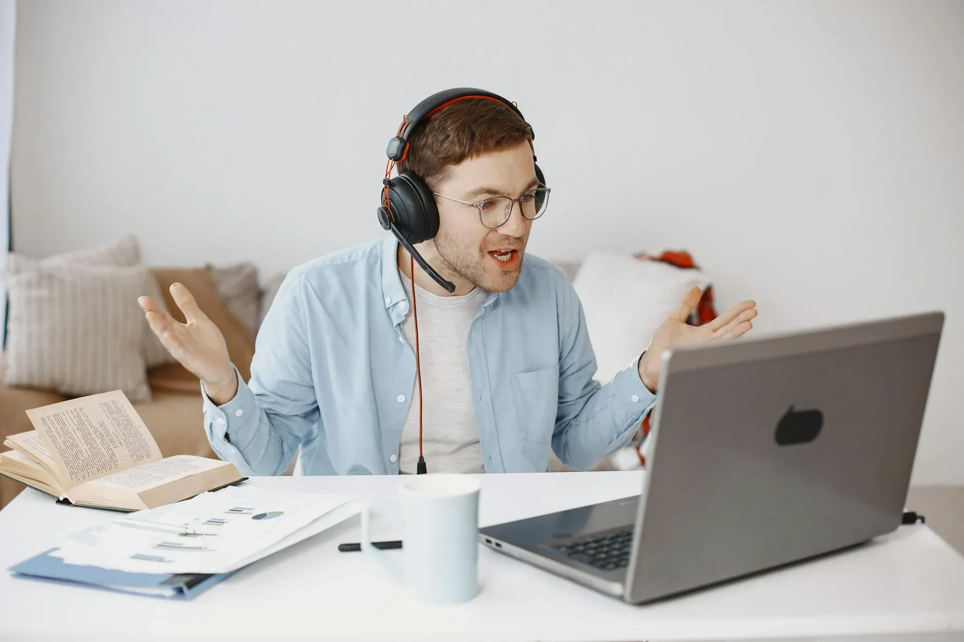 An office worker in a home setting appears frustrated during an online meeting, with a cluttered desk and laptop screen visible.