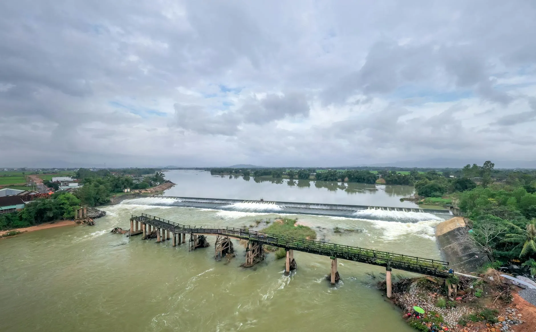 A drone shot capturing a damaged bridge spanning a river under a cloudy sky, highlighting structural issues and natural surroundings.