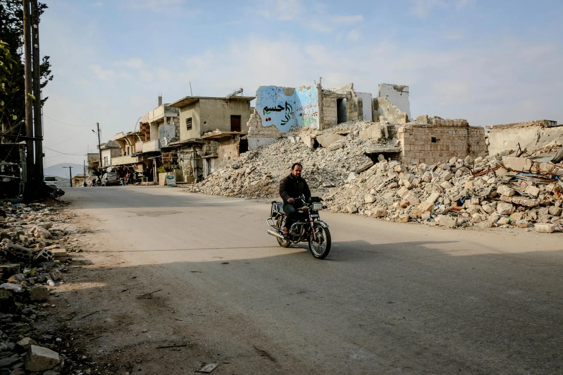 A lone rider on a motorcycle traverses a desolate road lined with destroyed buildings and rubble, symbolizing conflict's aftermath.
