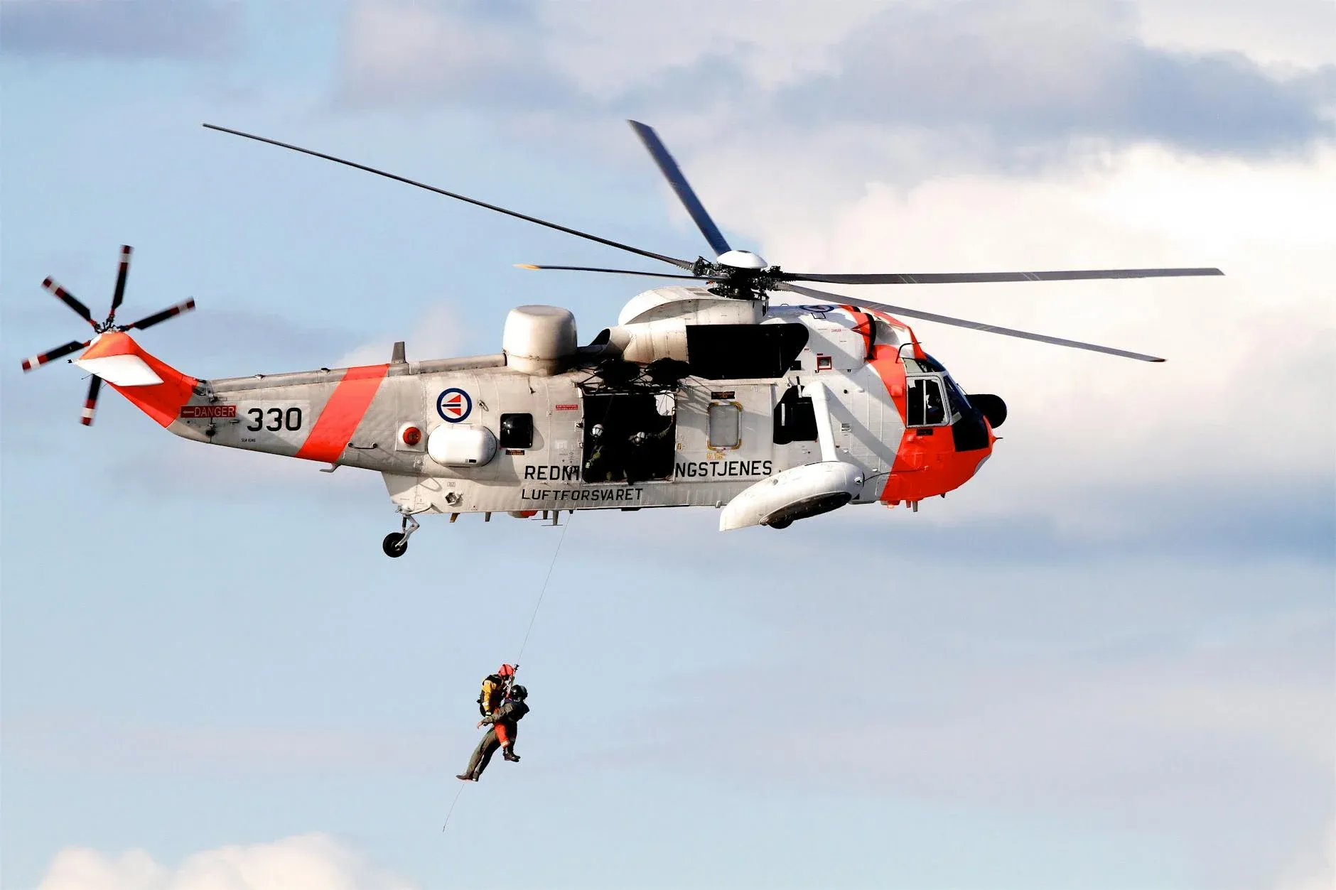 A rescue helicopter from the Royal Norwegian Air Force performs a mid-air hoist operation on a clear day.