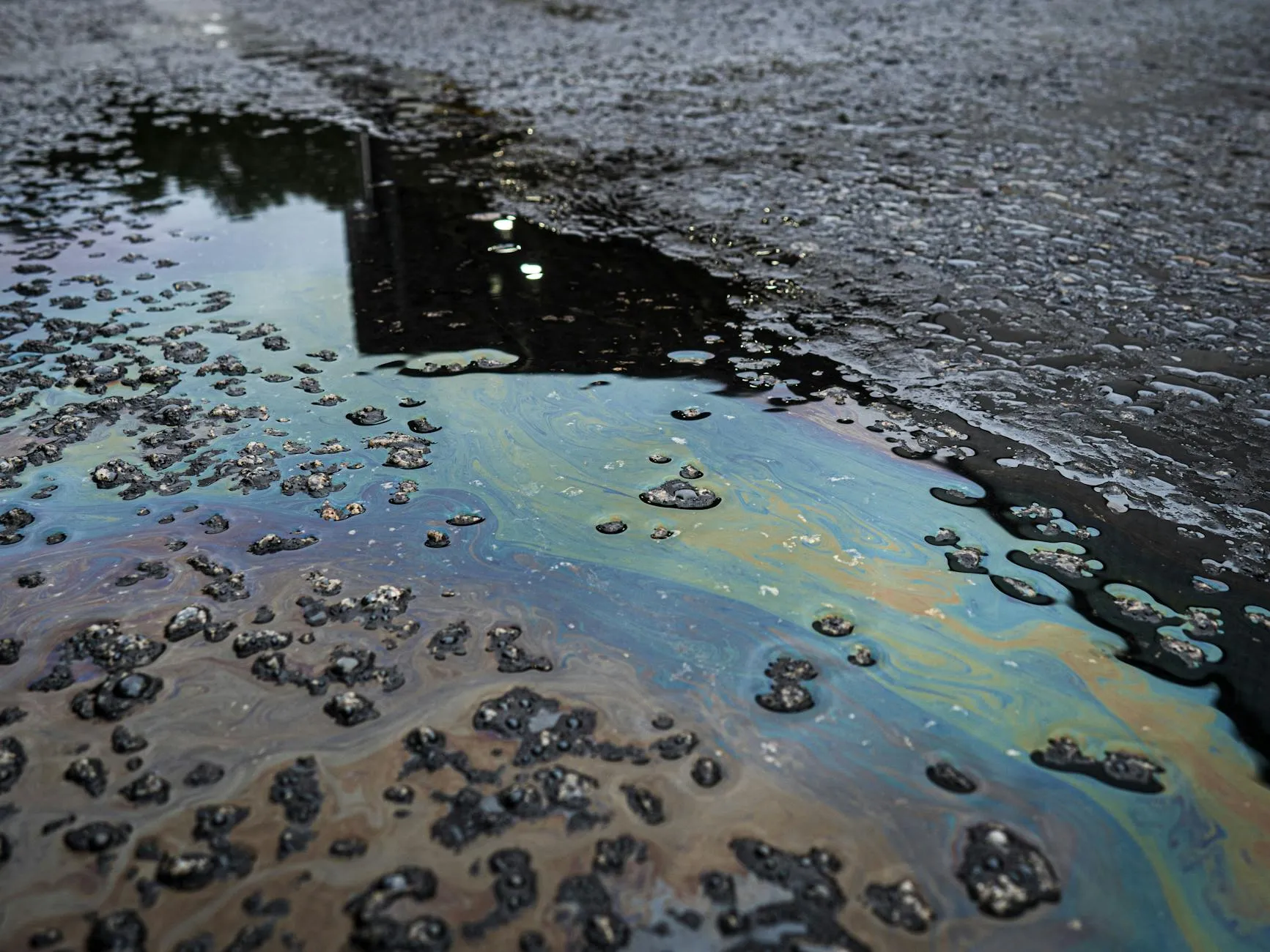 A vivid iridescent oil spill spreads across a rough asphalt surface, emphasizing environmental contamination.