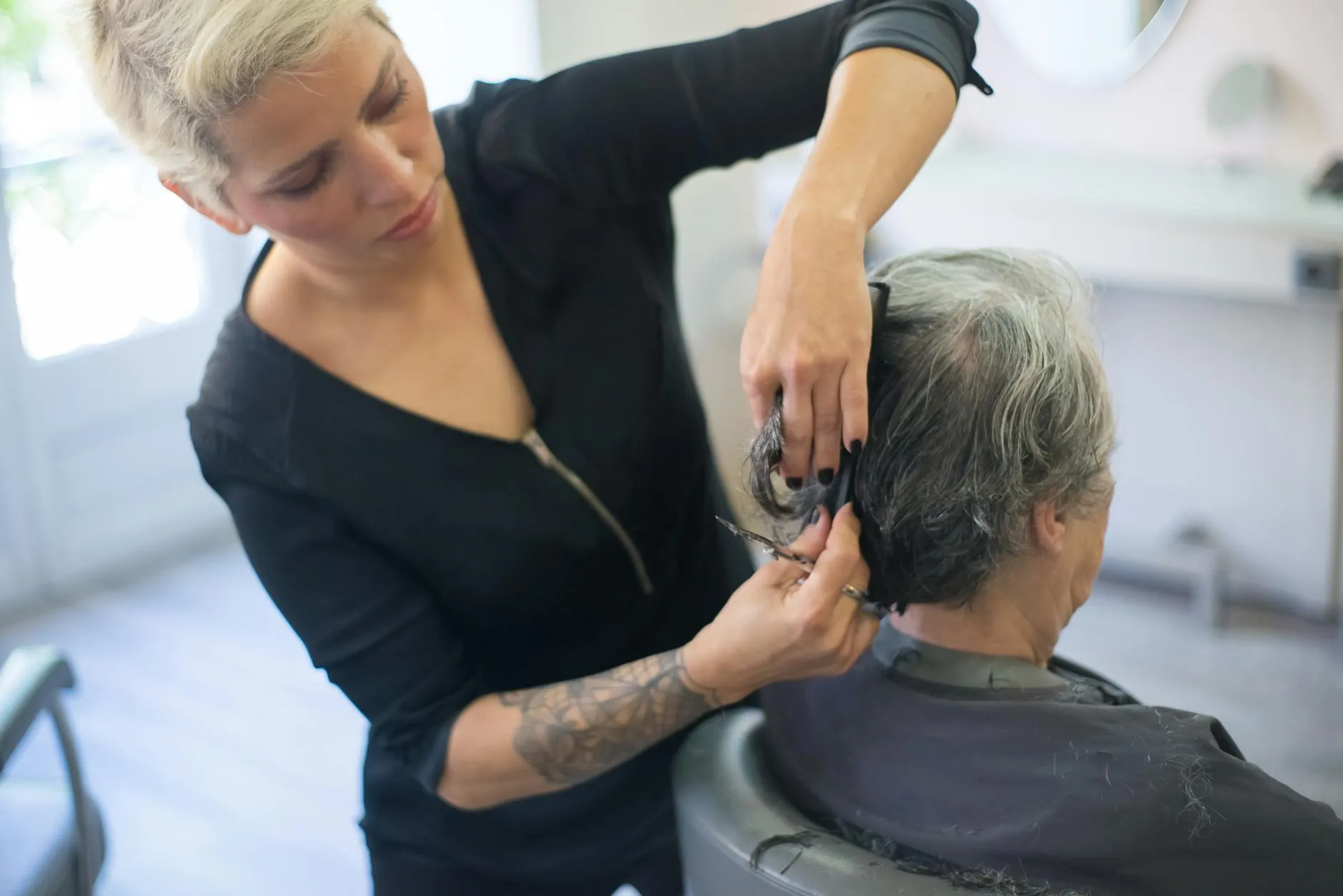 A skilled hair stylist with a tattoo is meticulously cutting a client's hair in a modern salon setting.