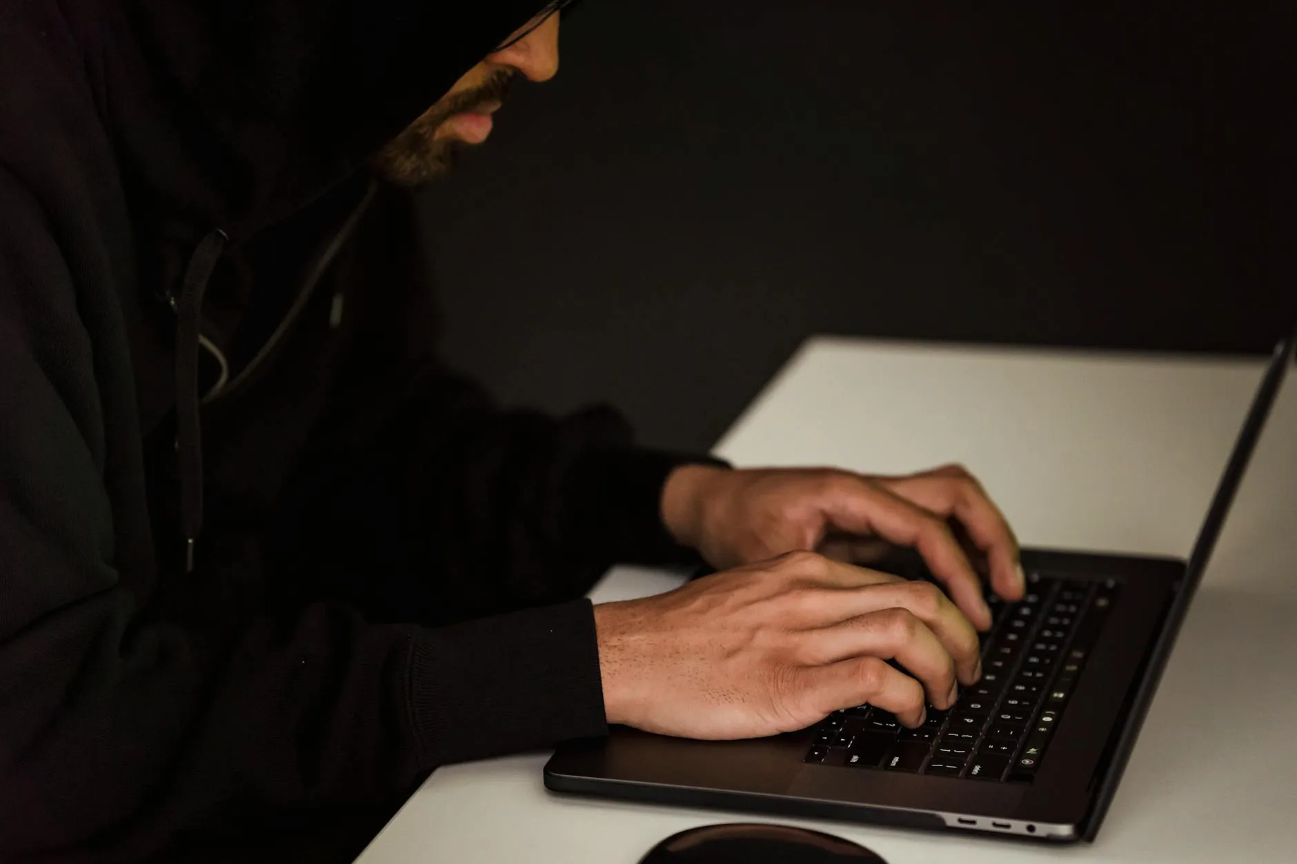Et skægget mandligt hacker-motiv i sort hætte sidder og skriver på en netbook i et mørkt, uoplyst rum. Billedet formidler en stemning af cyberaktivitet og digital efterforskning.