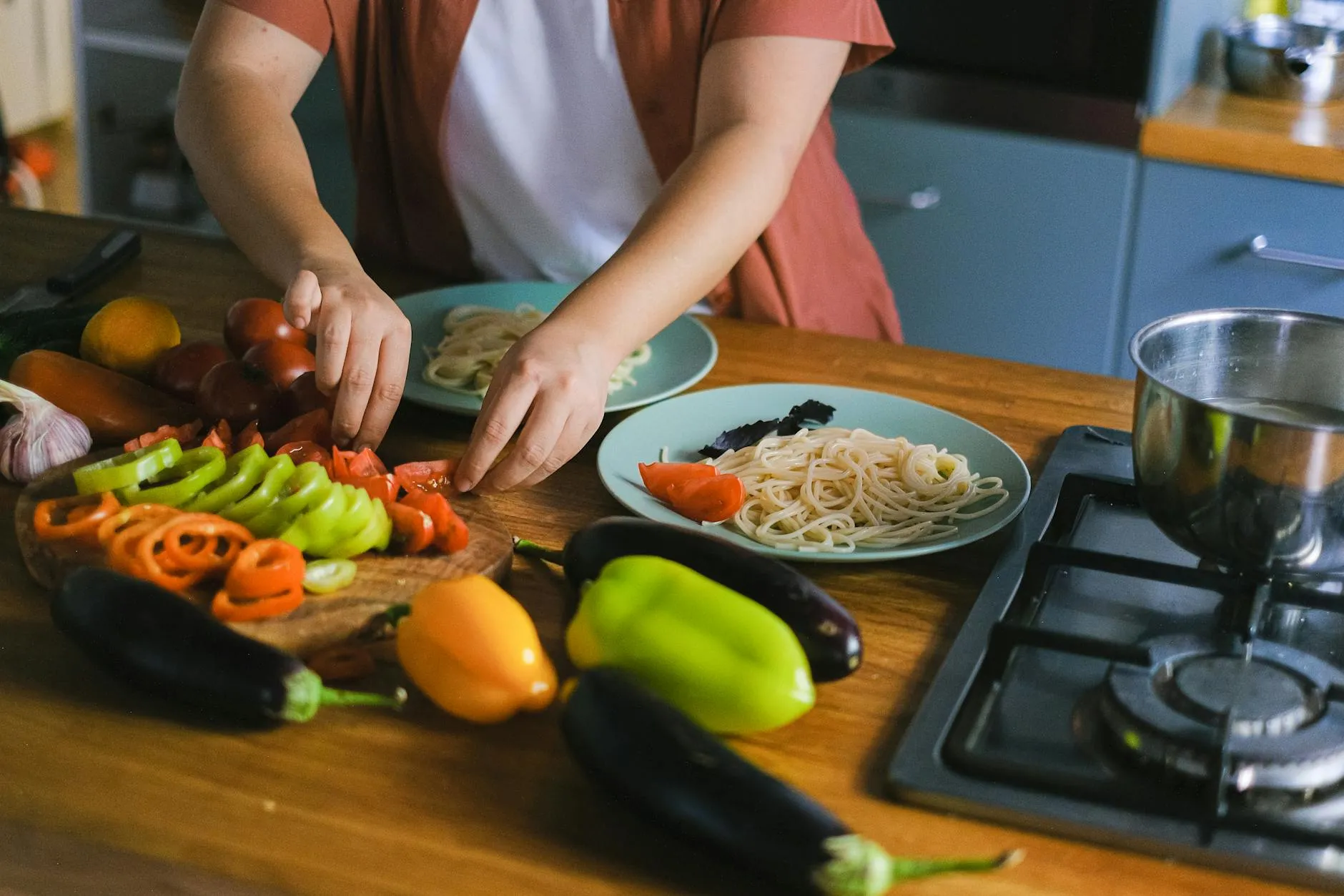 A woman carefully arranges colorful vegetables and pasta on plates in a well-lit kitchen, highlighting fresh ingredients.