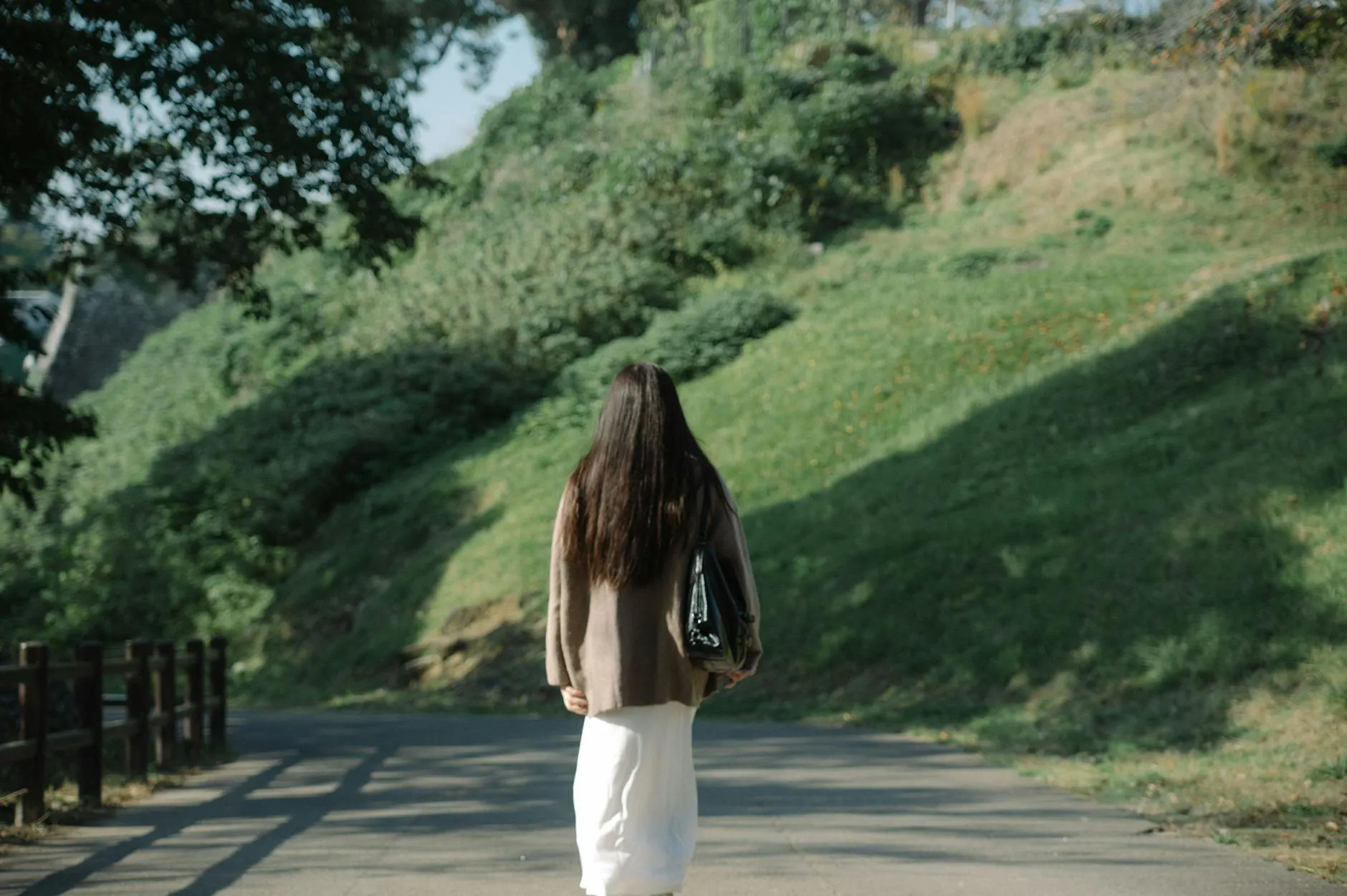 A serene scene of a woman walking alone on a sunlit path surrounded by lush greenery in a tranquil Japanese park.