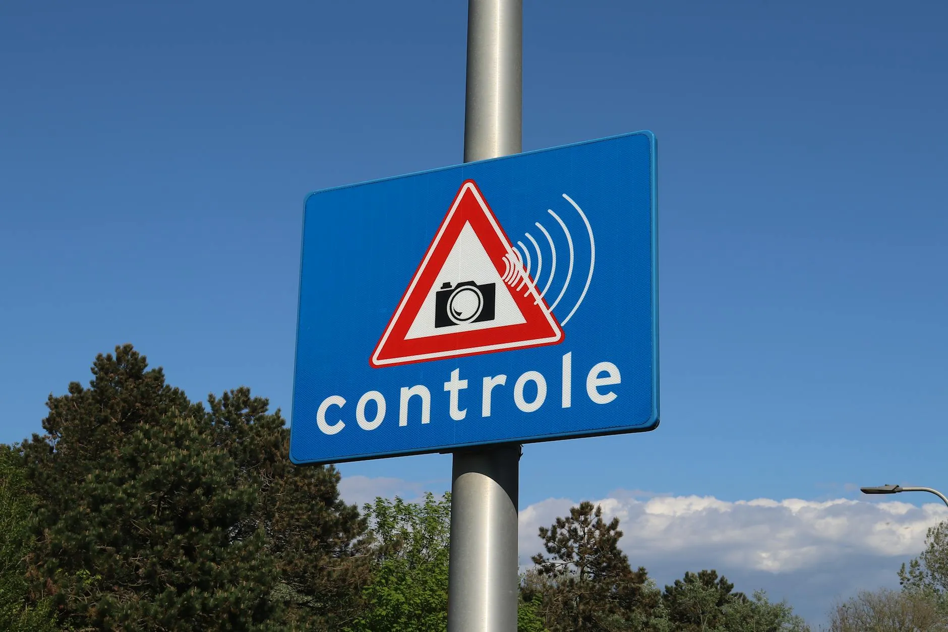 Et blåt vejskilt med et kamera-ikon og teksten “controle” markerer, at der foretages hastighedskontrol. Skiltet signalerer overvågning langs vejen.