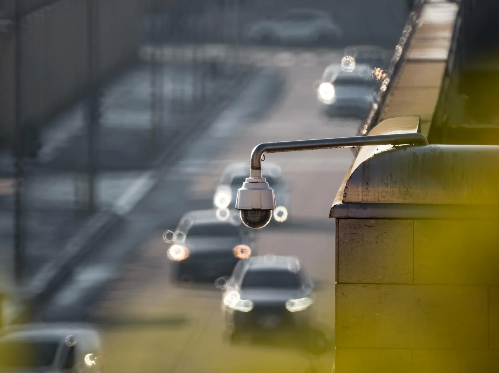 Et CCTV-kamera overvåger bytrafik på en travl vej. I baggrunden ses slørede køretøjer, der understreger bevægelse og overvågning i bymiljøet.