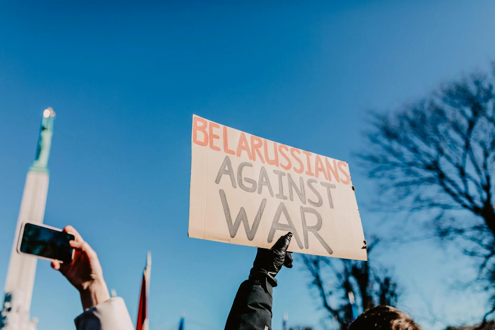 Demonstranter i Riga holder et skilt med teksten “Belarussians Against War” under en klar blå himmel. Billedet formidler en freds- og anti-krigsprotest.
