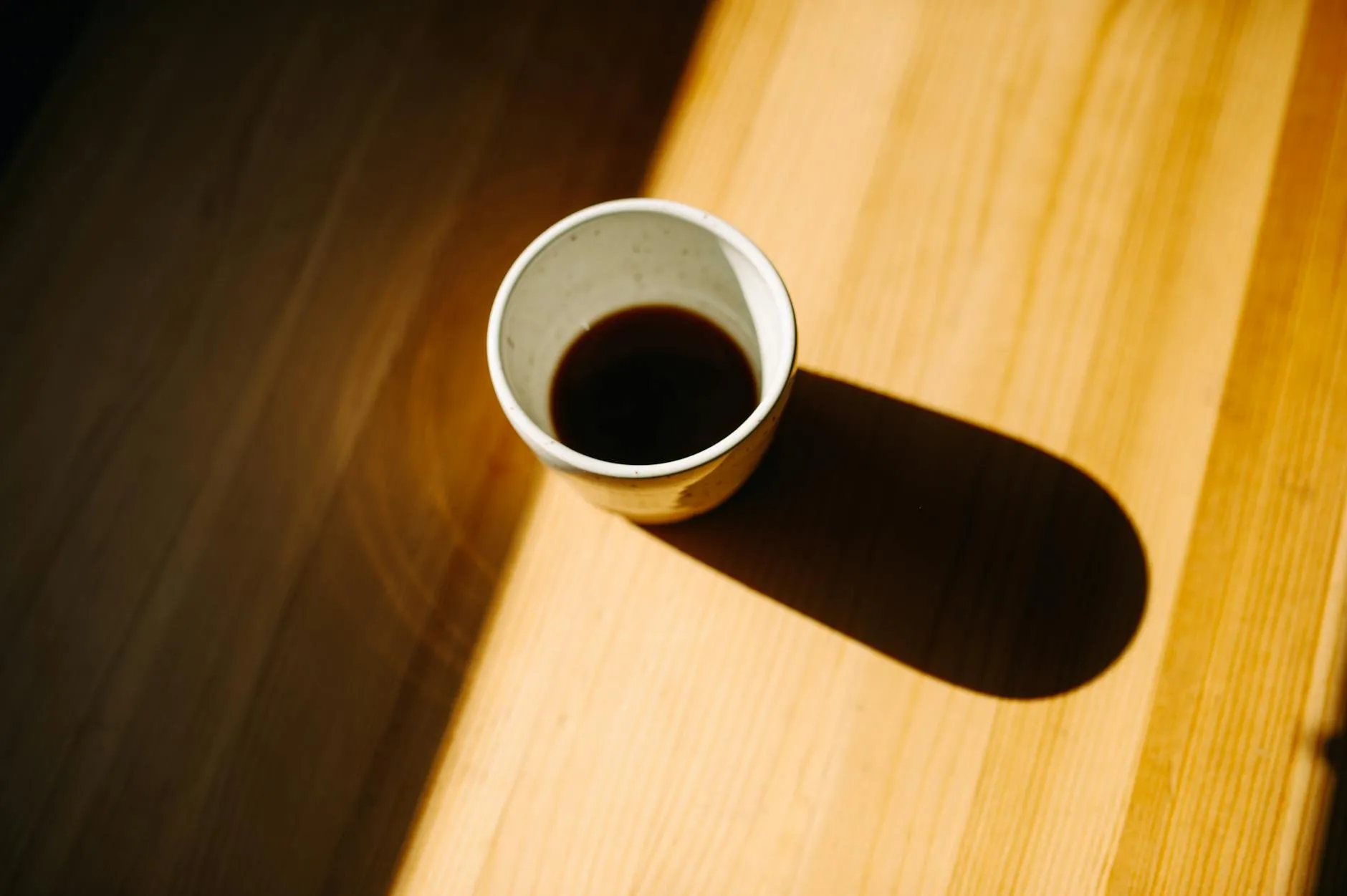 En kaffekop står på et træbord og kaster en tydelig skygge i det varme morgenlys. Scenen udstråler ro og hygge med et naturligt, indbydende udtryk.