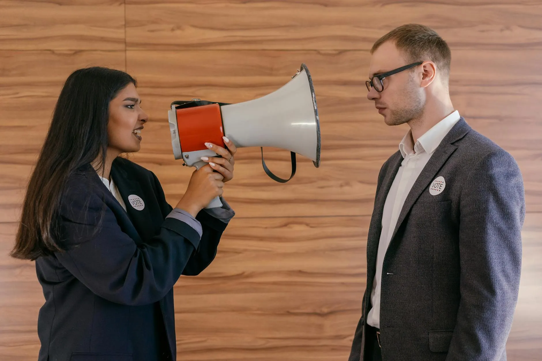 En kvinde bruger en megafon til at konfrontere en mand i jakkesæt indendørs. Motivet symboliserer politisk debat og uenighed.