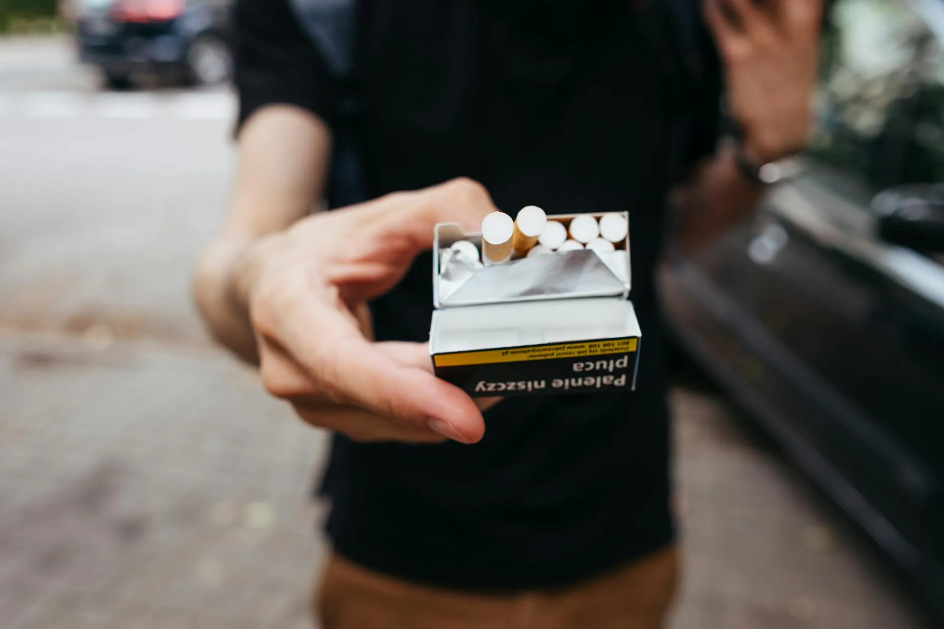 Et close-up af en mand, der rækker en pakke cigaretter frem udendørs på en bygade. Stemningen er hverdagsnær og dokumentarisk.