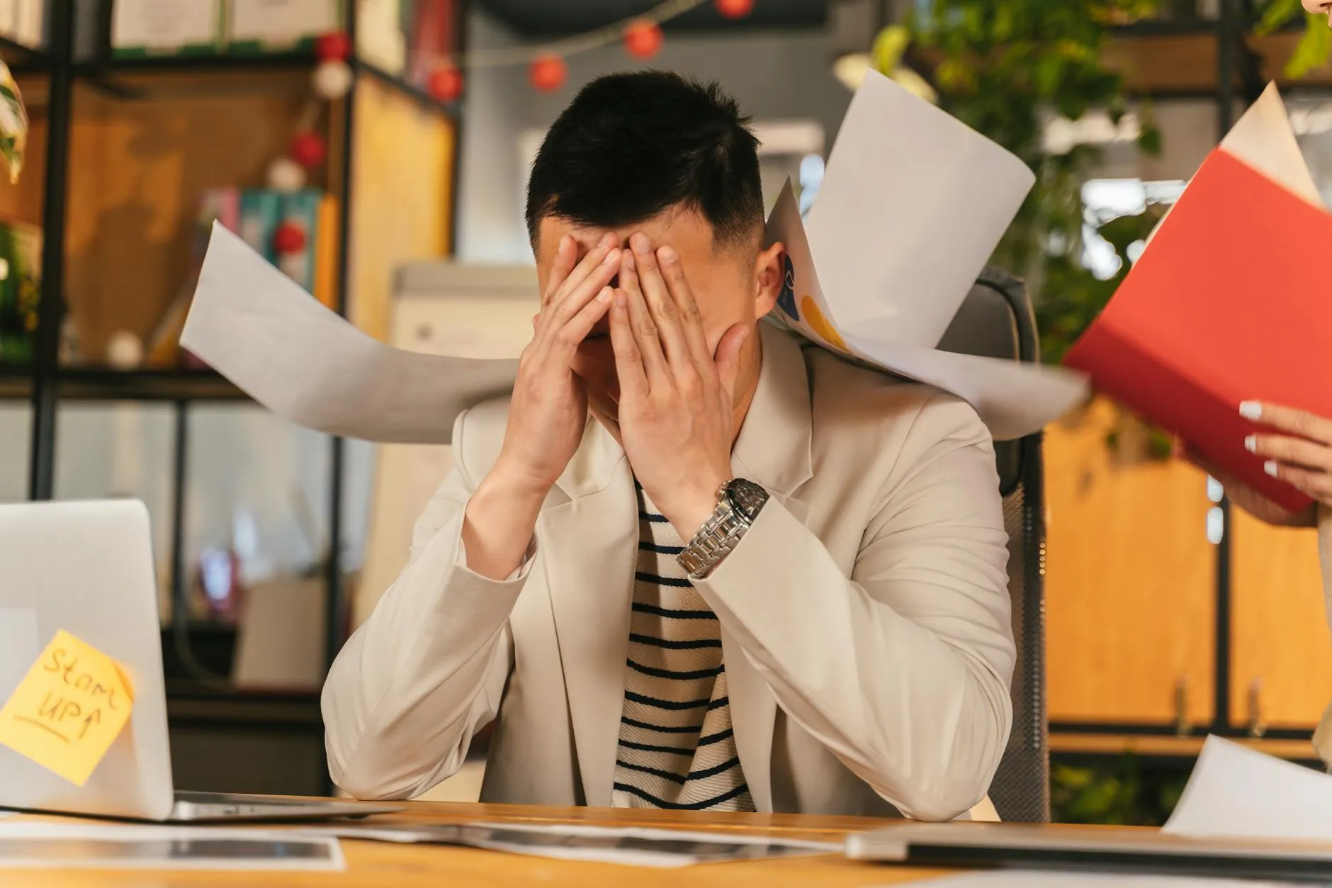 En stresset forretningsmand sidder i et kontormiljø omgivet af papirarbejde, der illustrerer arbejdsrelateret udmattelse. Billedet formidler tydeligt temaet burnout og pres i hverdagen.