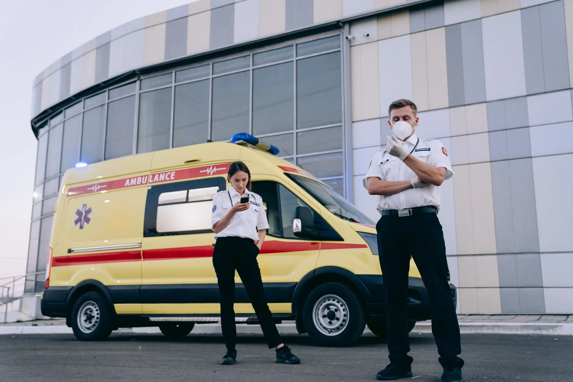 To paramedicinere i uniform står ved en ambulance uden for en moderne bygning. De er klar til at rykke ud og yde akut hjælp.
