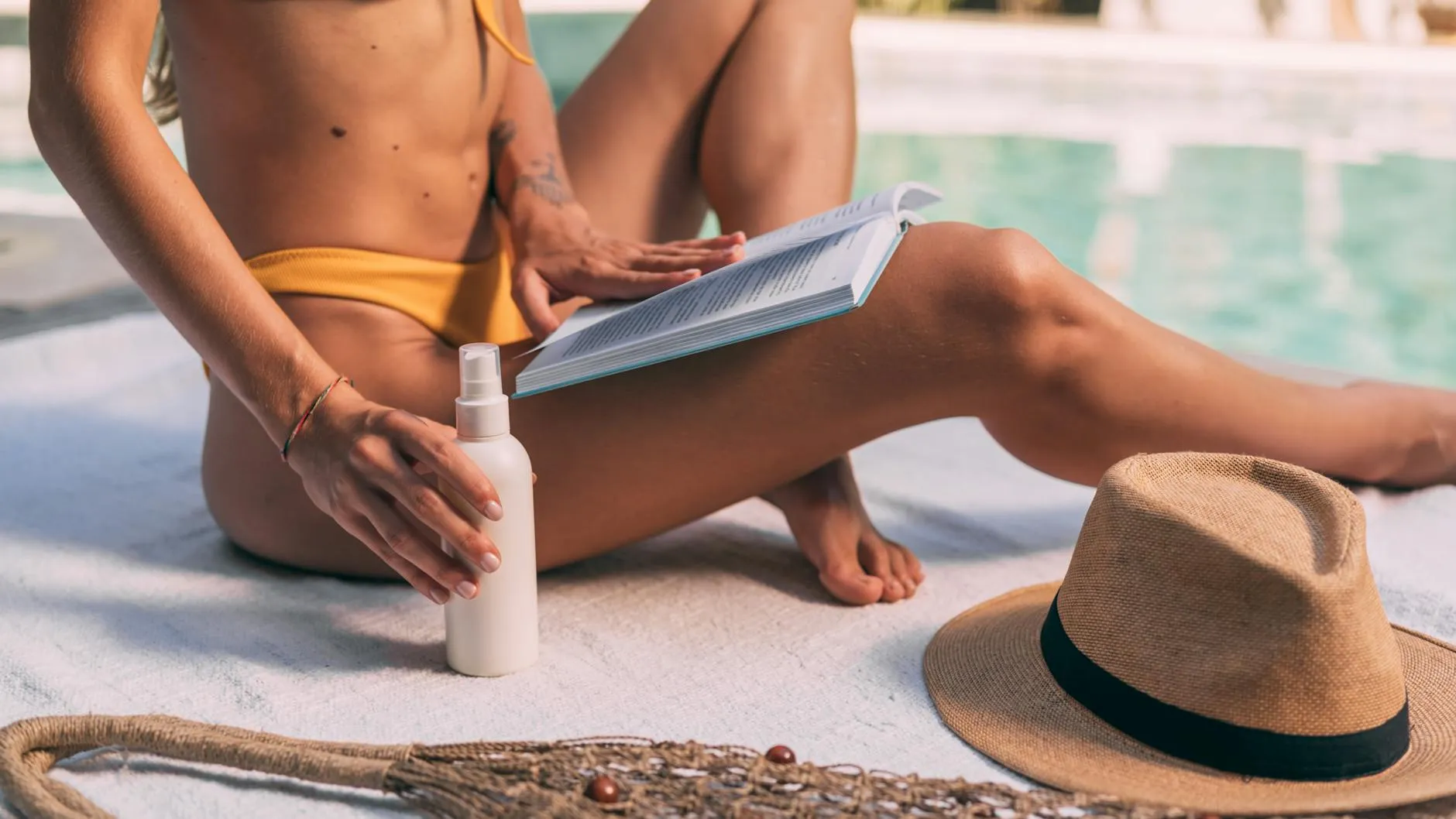 En kvinde i bikini læser ved poolen med solhat og solcreme, mens hun nyder sommerstemningen. Stemningen er afslappet og solrig.