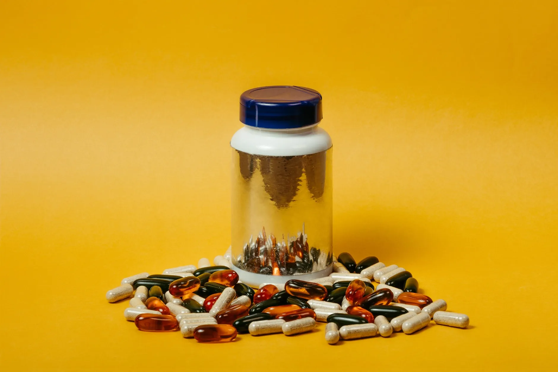 En samling af flerfarvede piller og kapsler ligger omkring en reflekterende pilleflaske på en lys gul baggrund. Billedet illustrerer medicin og farmaceutiske produkter.