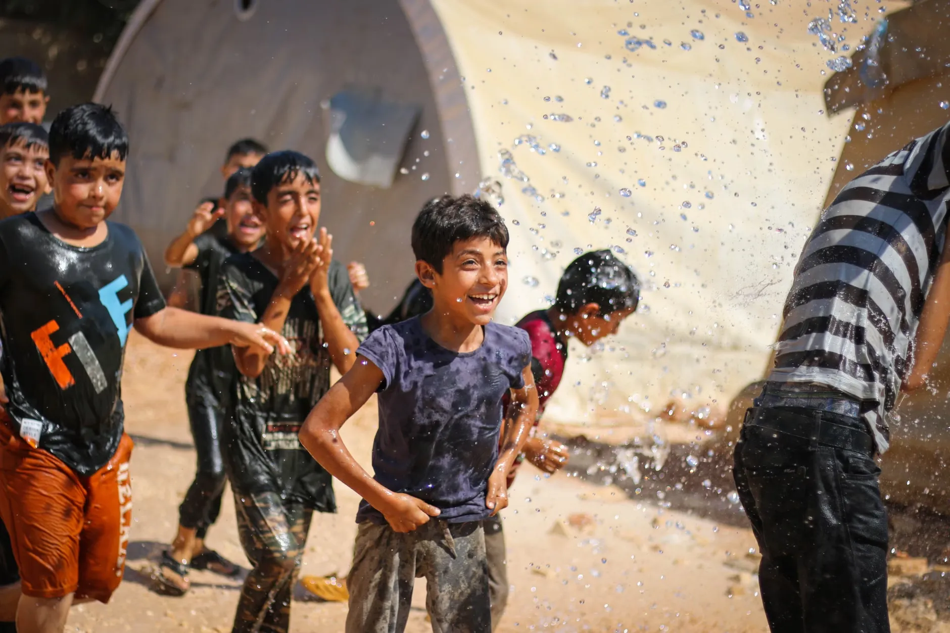 En gruppe glade børn leger og sprøjter vand på en solrig dag udendørs i Idlib, Syrien. Billedet fanger sommerglæde og leg.