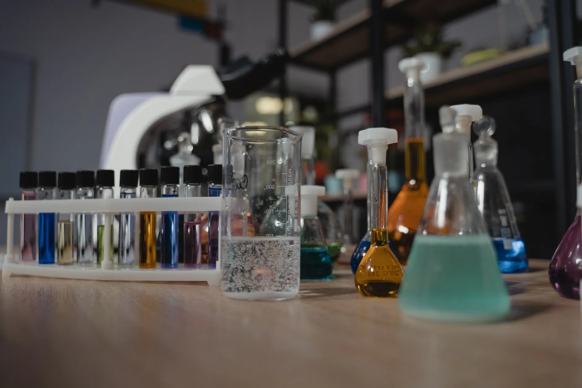 Laboratorieglas med farvede væsker står på et træbord, og billedet illustrerer kemisk forskning og forskellige laboratorieværktøjer. Scenen er fotograferet i et rent, oplyst miljø.