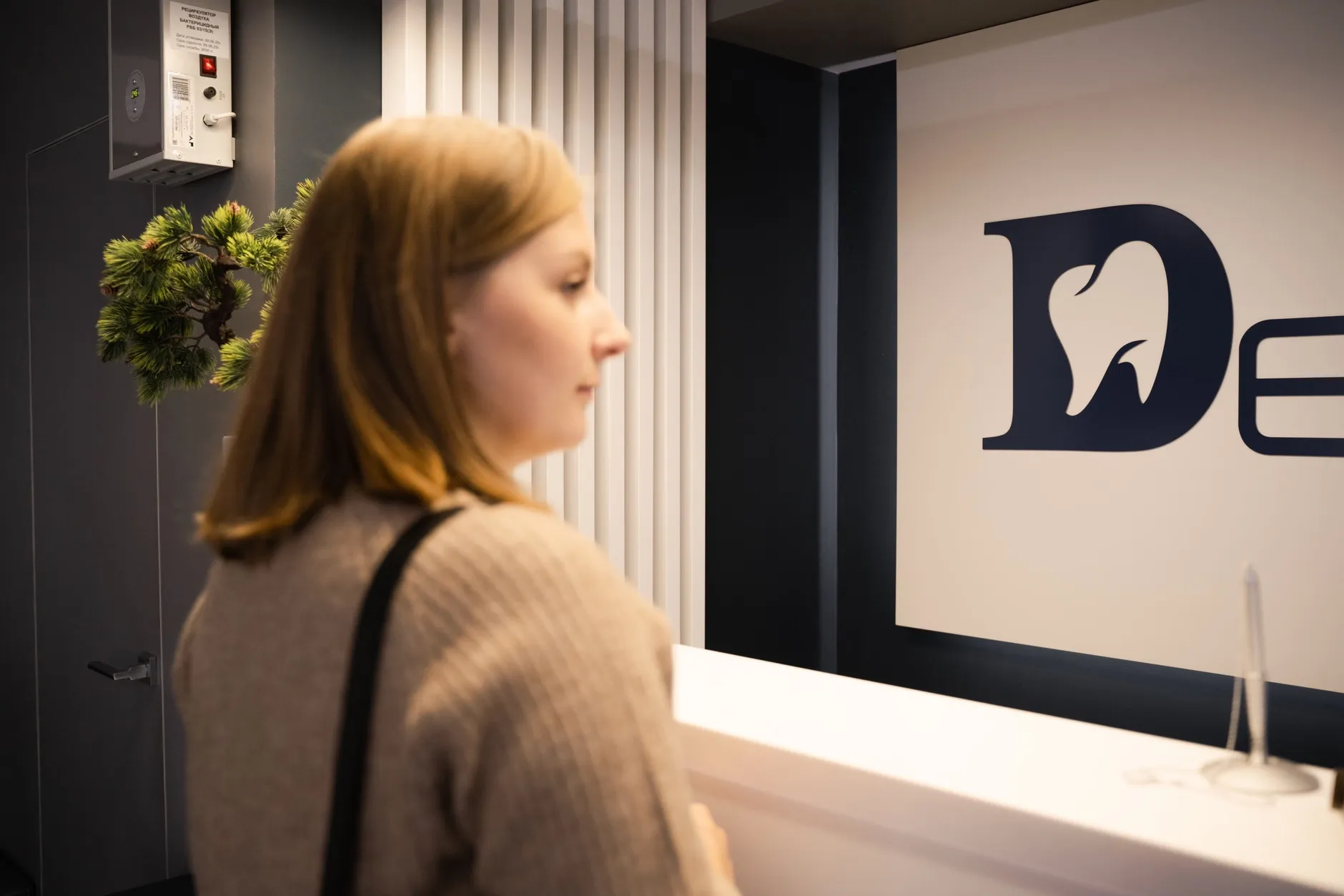 En ung kvinde står ved receptionen i en tandlægeklinik i sideprofil. Stemningen er rolig og hverdagsnær.