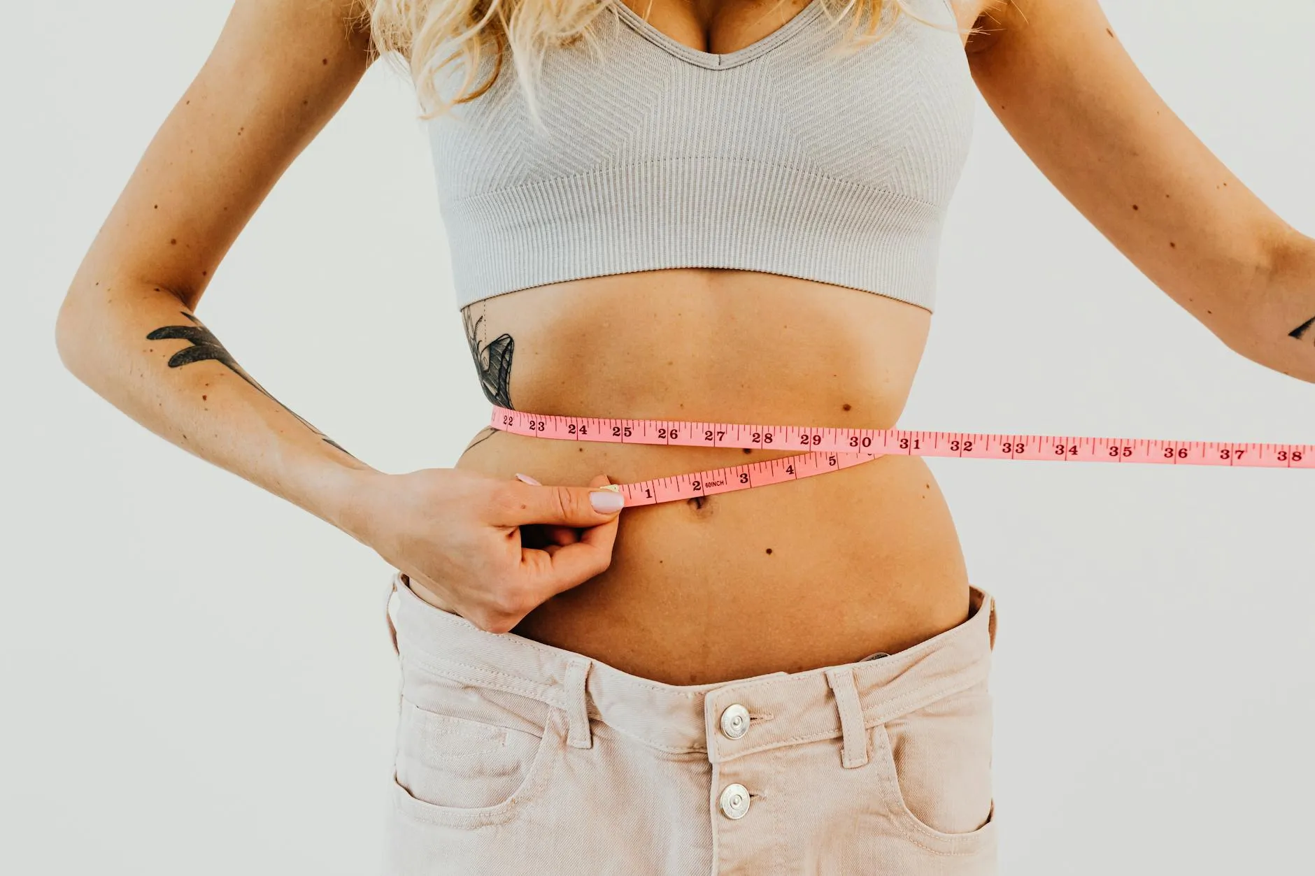 En kvinde måler sin talje med et pink målebånd i forbindelse med fitnessmål. Billedet er et tæt close-up, der fremhæver fokus på træning og velvære.