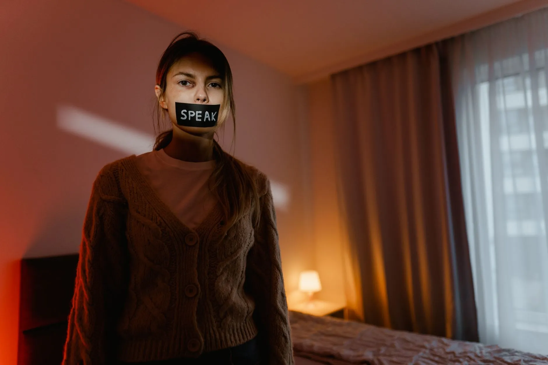 En kvinde står i et mørkt soveværelse med tape over munden med teksten “Speak”, som symboliserer tavshed og undertrykkelse. Stemningen er intens og understreger temaet om at blive holdt tilbage.