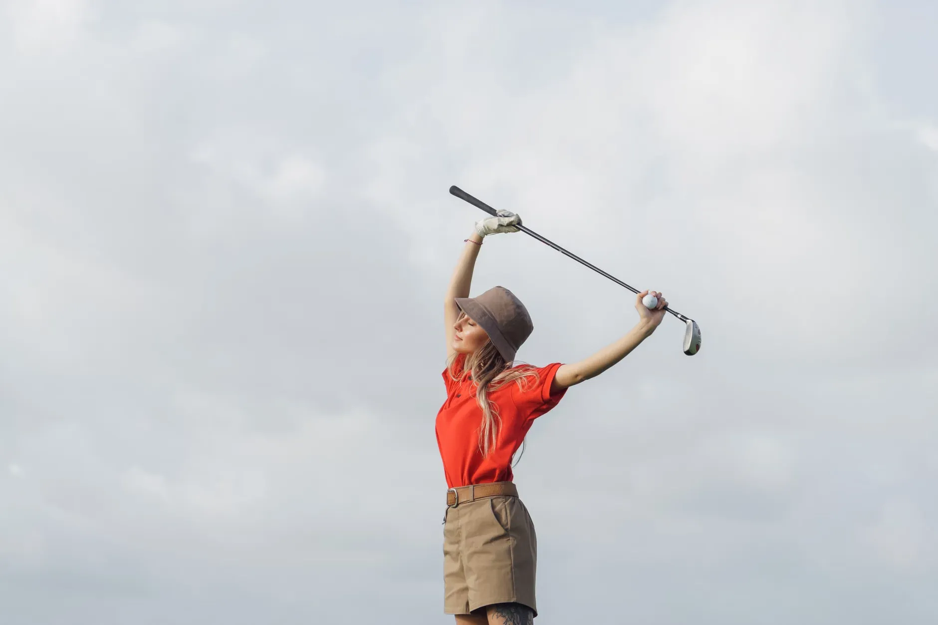 En kvinde i rød skjorte strækker sig med en golfkølle under skyede himmel, hvilket viser en aktiv livsstil. Billedet fremhæver motion og udendørsaktiviteter.