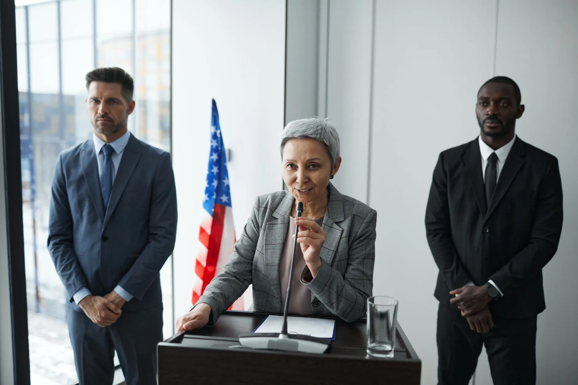 En kvindelig politiker holder en tale, mens livvagter står i baggrunden og et amerikansk flag ses. Billedet formidler politisk fremmøde og sikkerhed omkring arrangementet.
