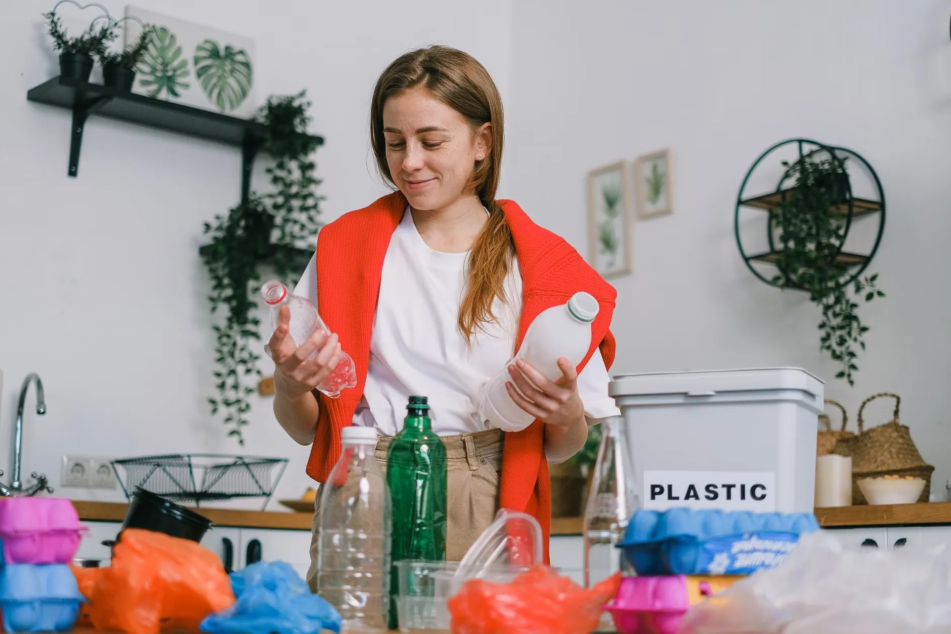 En glad kvinde i afslappet tøj står i et lyst køkken og sorterer affald med plastflasker i hænderne. Scenen er taget i dagslys og illustrerer genbrug og affaldssortering.
