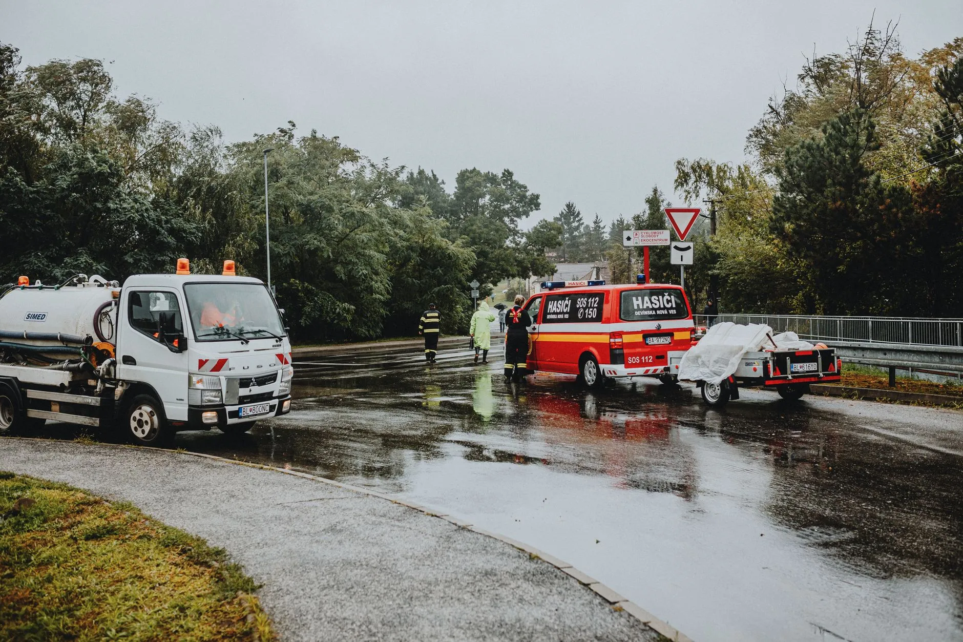 Redningspersonale og køretøjer er samlet ved en hændelse på en regnvåd vej. Scenen er fotograferet i dårligt vejr med synlige redningsindsatser.