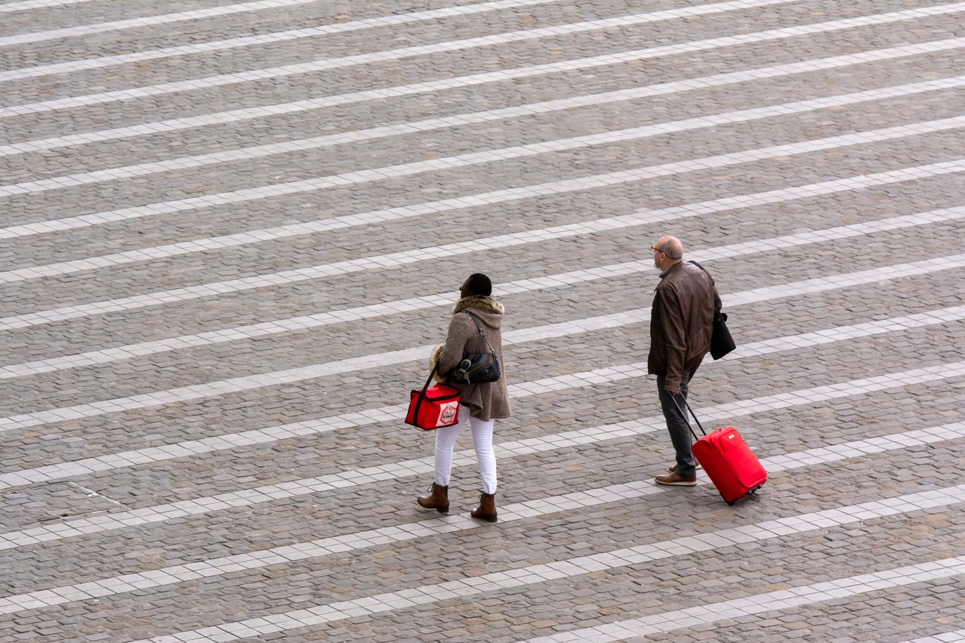 Et aerialt udsnit af mennesker, der går med kufferter hen over en mønstret bybelægning. Motivet understreger urban rejse og bevægelse.