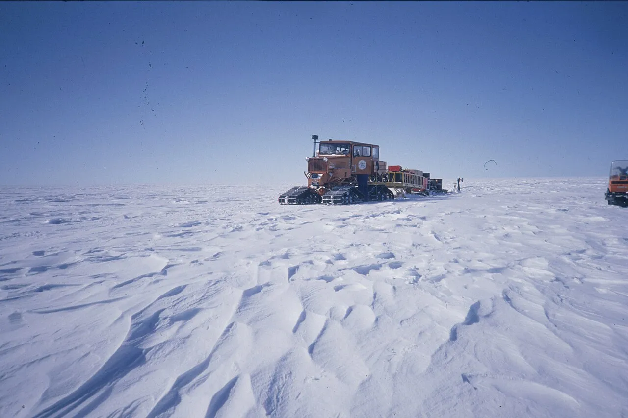 Små sastrugi-former ses på overfladen af Antarktis’ plateau, med en SnoCat-transport i baggrunden. Billedet illustrerer forholdene på den iskolde, vindformede slette.