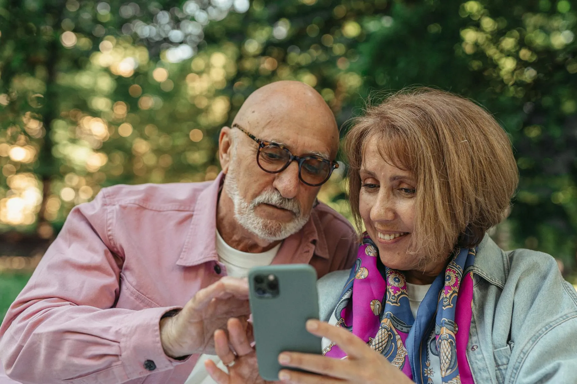 Et glad seniorpar sidder ude i en park og bruger en smartphone. Billedet formidler digital kommunikation og moderne hverdagsliv blandt ældre.