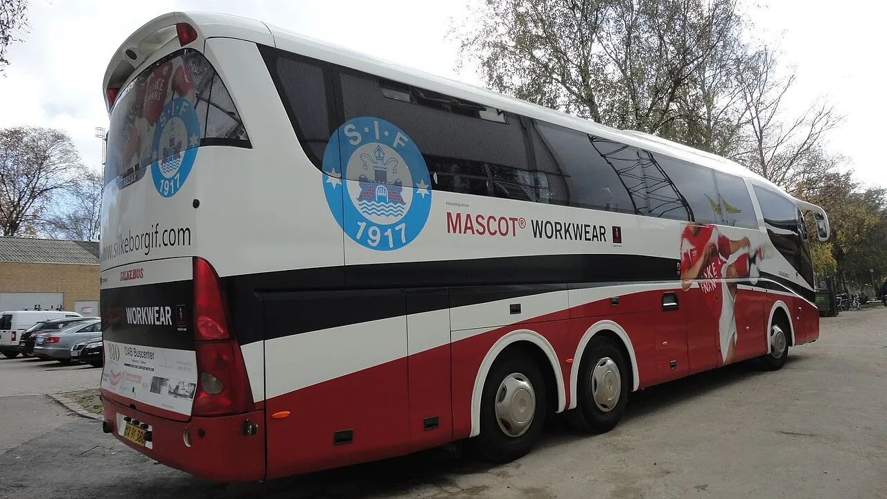 Holdbussen til fodboldklubben Silkeborg IF ses bagfra ved Lyngby Stadium på en overskyet dag. I bagenden og på siden ses klubbens logo samt sponsorinformation.