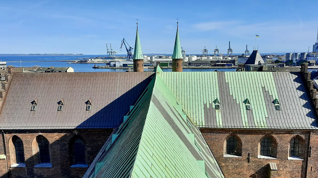 Udsigt fra Aarhus Domkirkes klokketårn hen over tagene mod havnens kraner og det dybblå hav. I baggrunden ses et ukrainsk flag ved den tidligere toldbygning.