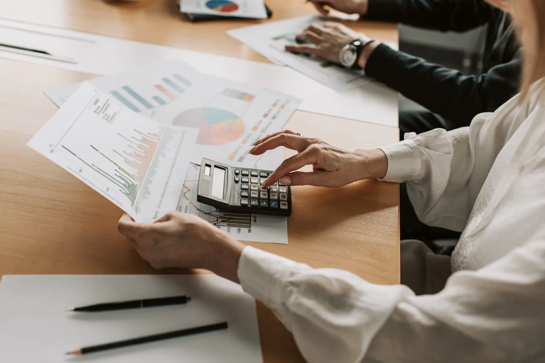 Hænder, der udfører finansielle beregninger med diagrammer og en lommeregner ved et mødebord. Billedet viser en person, der er dybt koncentreret omkring finansielle opgaver.