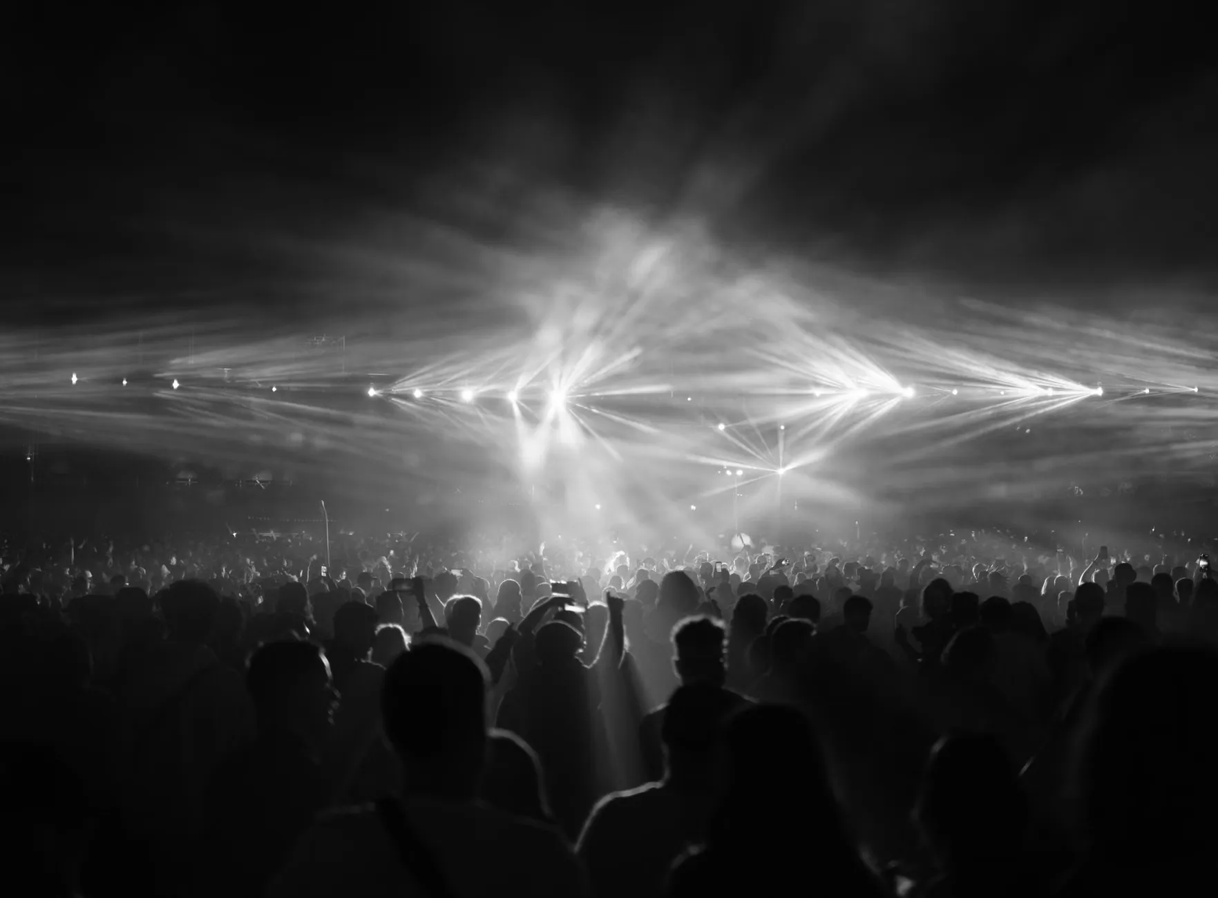 Et dramatisk sort-hvidt billede af et større publikum under en natkoncert med lysende scenelys.