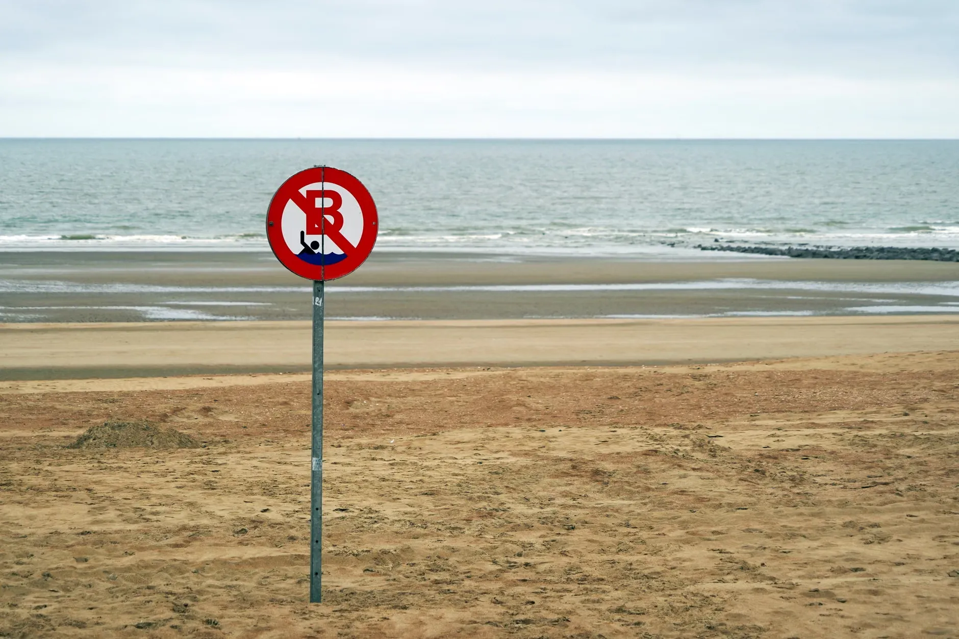 Et privat strandskilt, der viser regler for adgang, placeret ved en malerisk vandkant under en skyet himmel. Billedet viser en fredelig og rolig atmosfære.