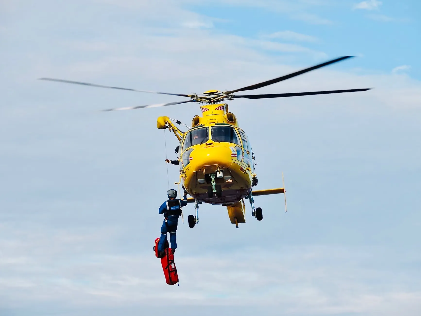 En gul helikopter udfører en redningsaktion med en besætningsmedlem hængende under.