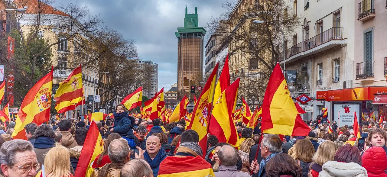 Billedet viser det spanske flag, et symbol på landets nationalidentitet og stolthed. Flaget er omgivet af mennesker i en livlig gade i Madrid.