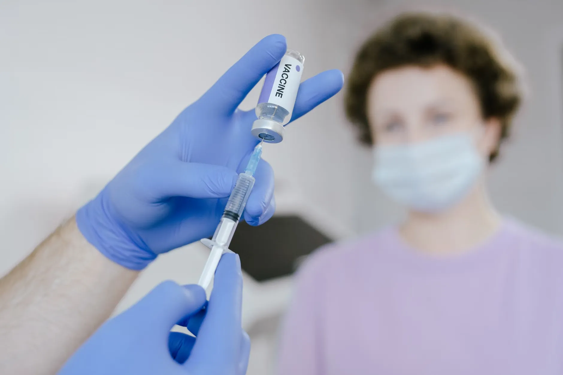 Et nærbillede af en sundhedsprofessionel i blå handsker, der fylder en sprøjte med vaccine. I baggrunden ses en patient, der venter på behandling.